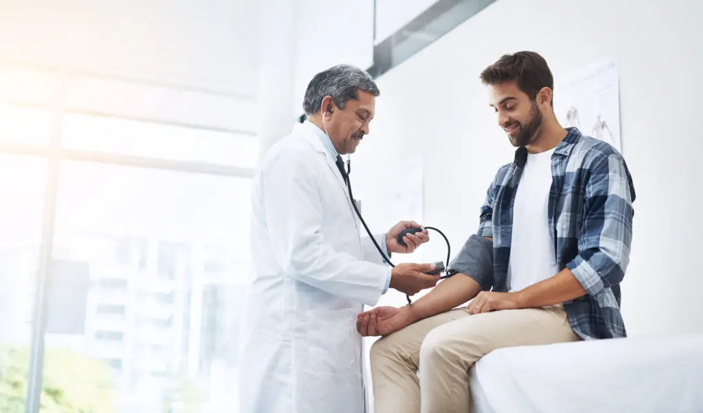 Healthcare, patient and doctor with blood pressure test, help and wellness checkup of person at hospital. Medicine, health care expert and Indian man in doctors office consulting medical professional